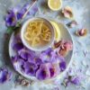 A refreshing beverage in a rustic cup with crystallized ginger on top, accented by a lemon slice and a sprig of mint, surrounded by an array of purple pansies and rose petals on a textured surface.