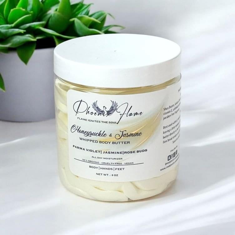 A jar of Phoenix Flame Whipped Body Butter with a label featuring essential ingredients like Parma Violet, Jasmine, and Rose Buds, placed near a green plant on a soft white surface.